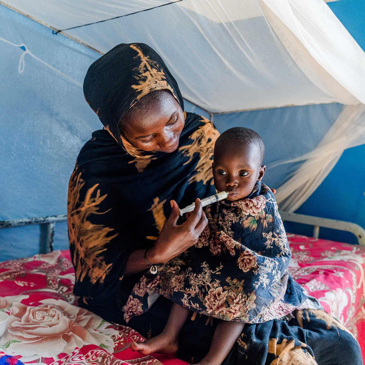 A mother helps her young child take medicine with a syringe while sitting together on a bed under a mosquito net.